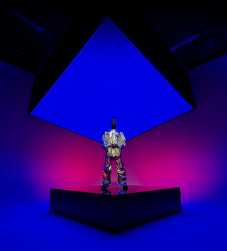 Imagem de uma pessoa vestida com uma roupa metálica em pé em um palco quadrado que se estende até o fundo, capturada na câmera ultra-angular do iPhone 16 Pro Max