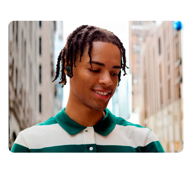 Vemos um homem usando Galaxy Buds3 FE pretos ao ar livre, com a cidade ao fundo, utilizando o recurso de chamadas nítidas para melhorar a qualidade da chamada.