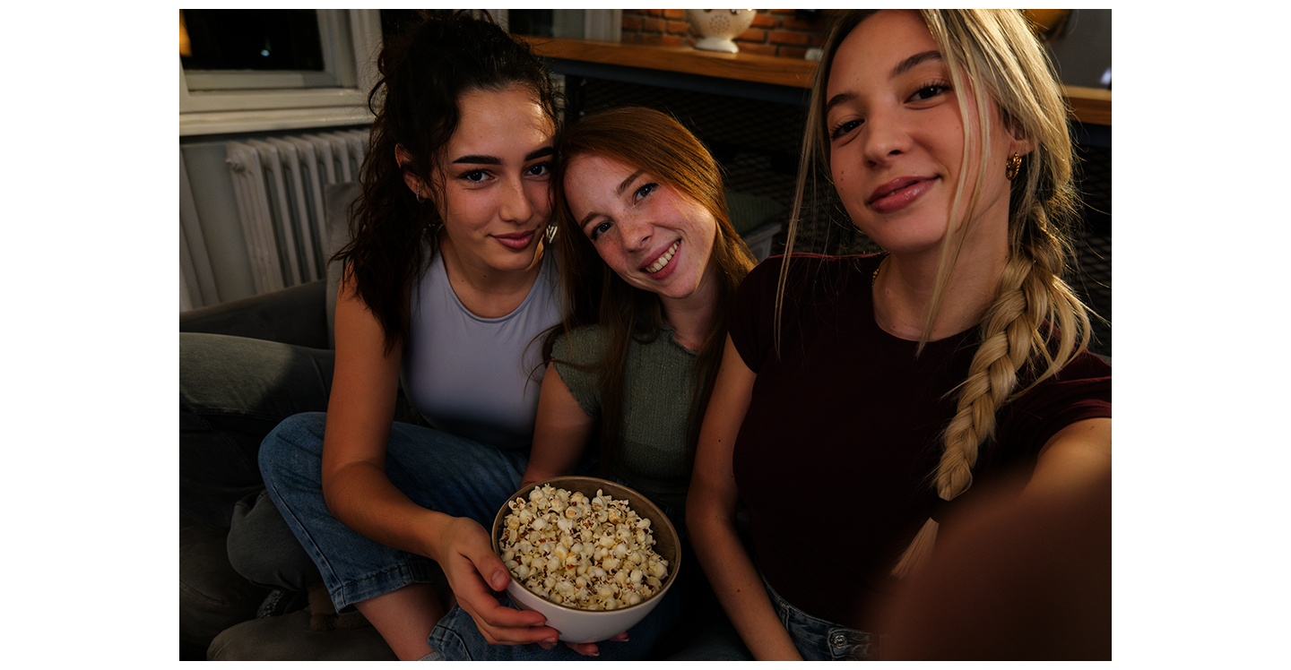 Três jovens estão sentadas em um sofá em casa, em um ambiente com pouca luz, tirando uma foto juntas. Uma delas tira a foto usando o Galaxy A37 5G com Nightography para registrar detalhes mais brilhantes e vibrantes.