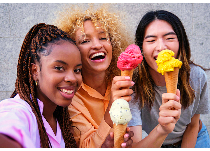 Três jovens, em pé e bem próximas, estão sorrindo e segurando casquinhas de sorvete enquanto tiram uma selfie. Uma delas tira a selfie usando a Câmera Frontal do Galaxy A37 5G para capturar detalhes vibrantes e nítidos.