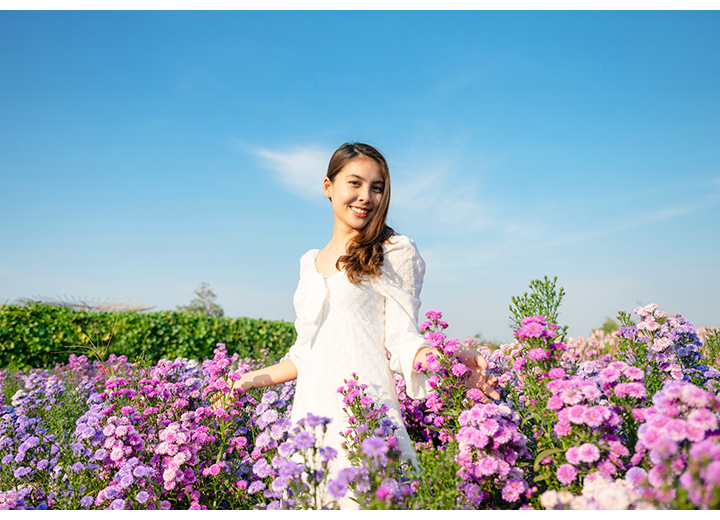 Uma mulher de vestido branco está em um ambiente externo, entre várias flores, posando para a foto.