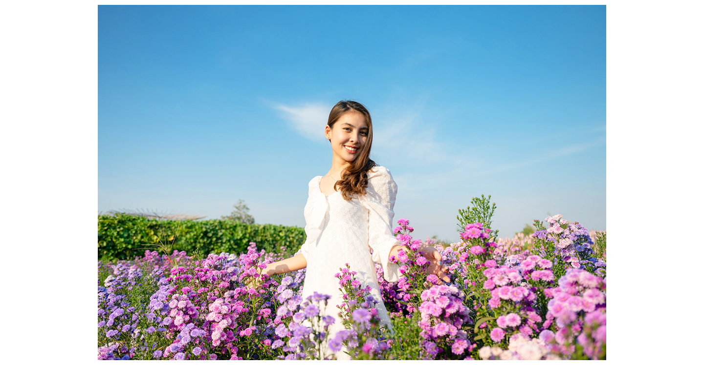 Uma mulher de vestido branco está em um ambiente externo, entre várias flores, posando para a foto.