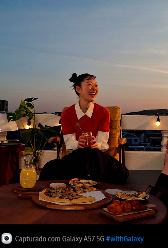Foto de uma jovem sorrindo em um terraço ao ar livre em ambiente com pouca luz, capturada com o Galaxy A57 5G. Os detalhes da foto aparecem claros e nítidos. Na parte inferior, aparece um ícone de câmera com o texto 'Capturado com Galaxy A57 5G' e a hashtag #withGalaxy.