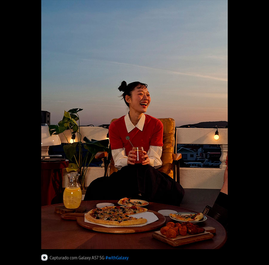 Foto de uma jovem sorrindo em um terraço ao ar livre em ambiente com pouca luz, capturada com o Galaxy A57 5G. Os detalhes da foto aparecem claros e nítidos. Na parte inferior, aparece um ícone de câmera com o texto 'Capturado com Galaxy A57 5G' e a hashtag #withGalaxy.