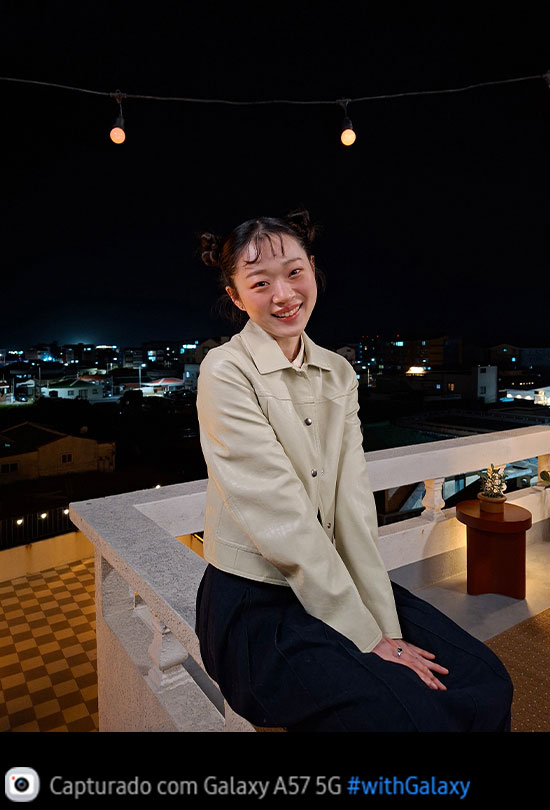 Foto de uma jovem apoiada em um terraço ao ar livre durante a noite, capturada com o Galaxy A57 5G. Os detalhes da foto aparecem claros e definidos graças ao Nightography otimizado. Na parte inferior, aparece um ícone de câmera com o texto 'Capturado com Galaxy A57 5G' e a hashtag #withGalaxy.