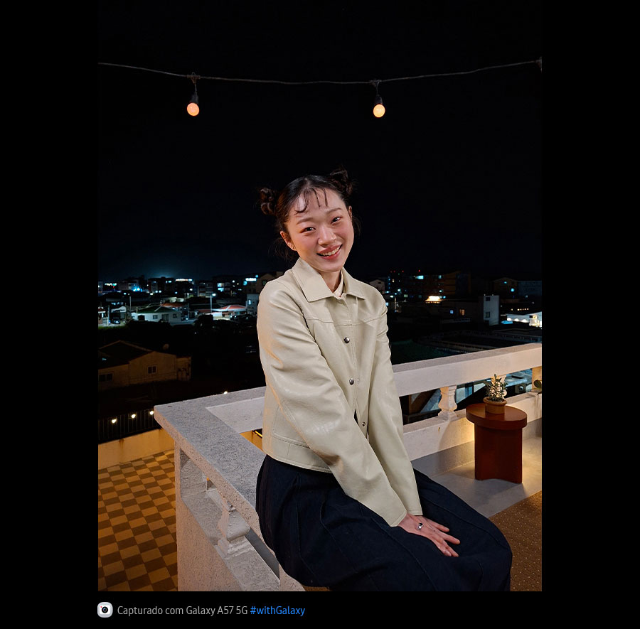 Foto de uma jovem apoiada em um terraço ao ar livre durante a noite, capturada com o Galaxy A57 5G. Os detalhes da foto aparecem claros e definidos graças ao Nightography otimizado. Na parte inferior, aparece um ícone de câmera com o texto 'Capturado com Galaxy A57 5G' e a hashtag #withGalaxy.