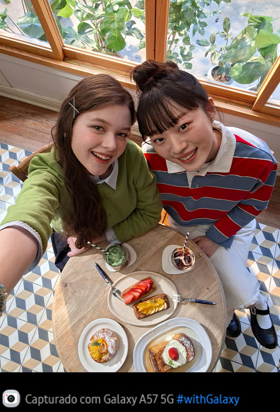 Foto selfie de duas jovens tomando brunch em um café bem iluminado, capturada de cima com a Câmera Frontal do Galaxy A57 5G. Os detalhes da foto são vibrantes e coloridos graças ao Super HDR. Na parte inferior, aparece um ícone de câmera com o texto 'Capturado com Galaxy A57 5G' e a hashtag #withGalaxy.