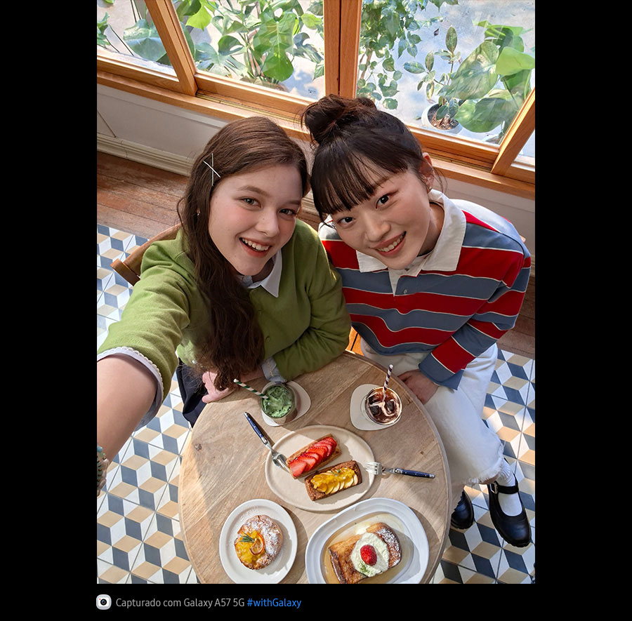 Foto selfie de duas jovens tomando brunch em um café bem iluminado, capturada de cima com a Câmera Frontal do Galaxy A57 5G. Os detalhes da foto são vibrantes e coloridos graças ao Super HDR. Na parte inferior, aparece um ícone de câmera com o texto 'Capturado com Galaxy A57 5G' e a hashtag #withGalaxy.