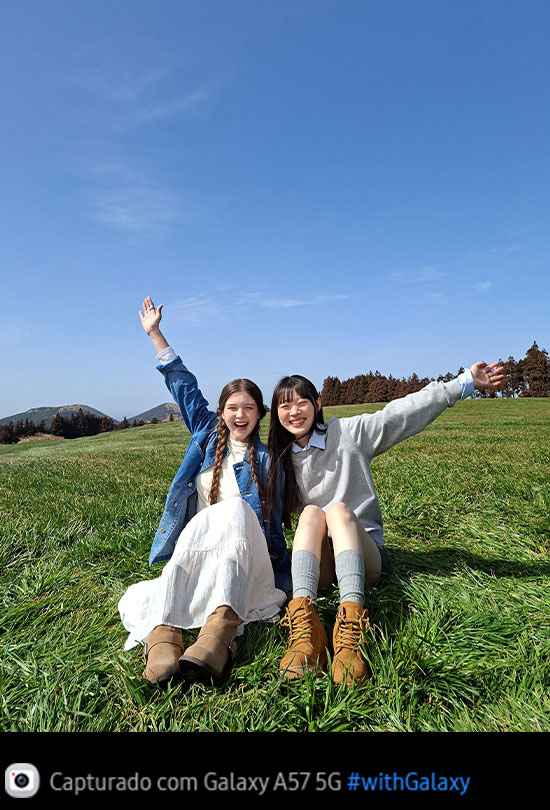Foto de duas jovens sentadas em um campo gramado aberto sob um céu azul, capturada com a Câmera Principal do Galaxy A57 5G. Os detalhes da foto são brilhantes e vívidos com qualidade aprimorada. Na parte inferior, aparece um ícone de câmera com o texto 'Capturado com Galaxy A57 5G' e a hashtag #withGalaxy.