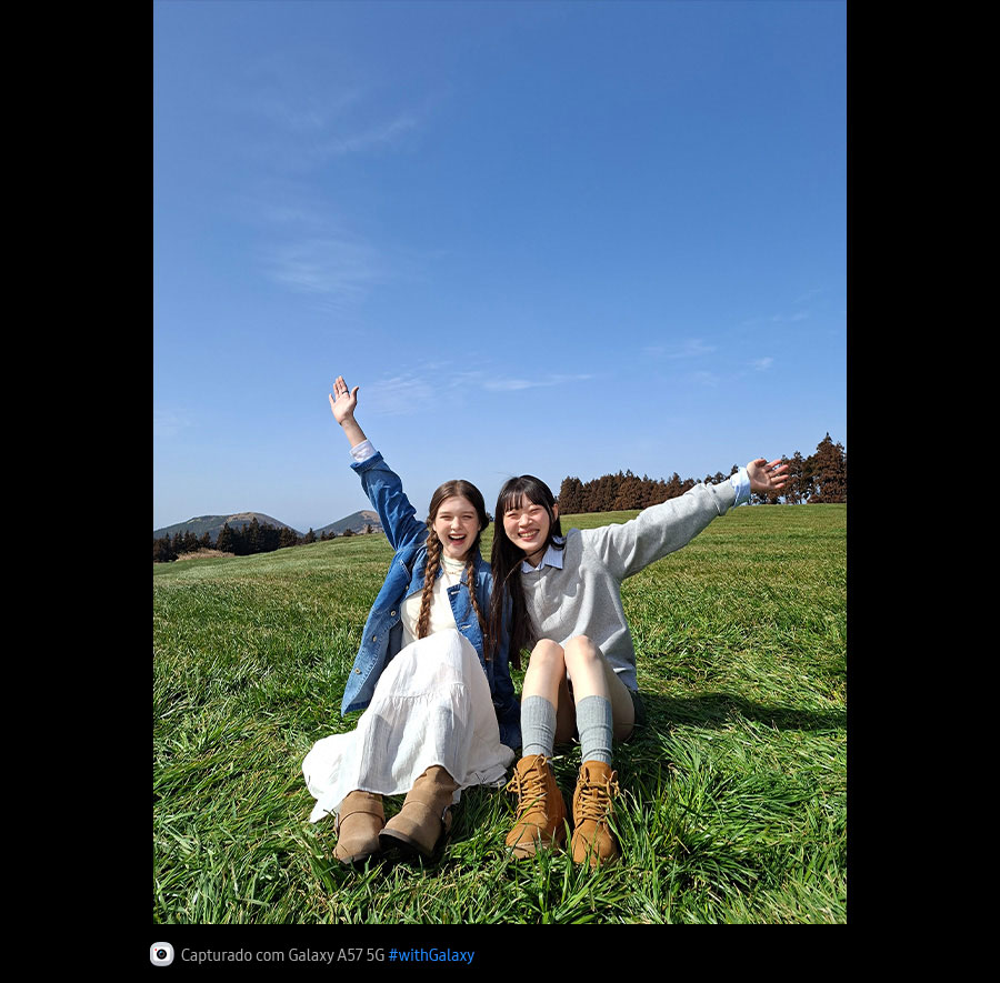 Foto de duas jovens sentadas em um campo gramado aberto sob um céu azul, capturada com a Câmera Principal do Galaxy A57 5G. Os detalhes da foto são brilhantes e vívidos com qualidade aprimorada. Na parte inferior, aparece um ícone de câmera com o texto 'Capturado com Galaxy A57 5G' e a hashtag #withGalaxy.