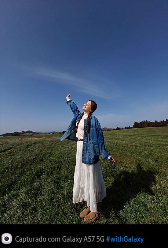 Foto de uma jovem em pé no meio de um campo gramado aberto sob um céu azul, capturada com a Câmera Ultra-Wide do Galaxy A57 5G. Os detalhes da imagem são claros e vívidos, com qualidade aprimorada. Na parte inferior, aparece um ícone de câmera com o texto 'Capturado com Galaxy A57 5G' e a hashtag #withGalaxy.