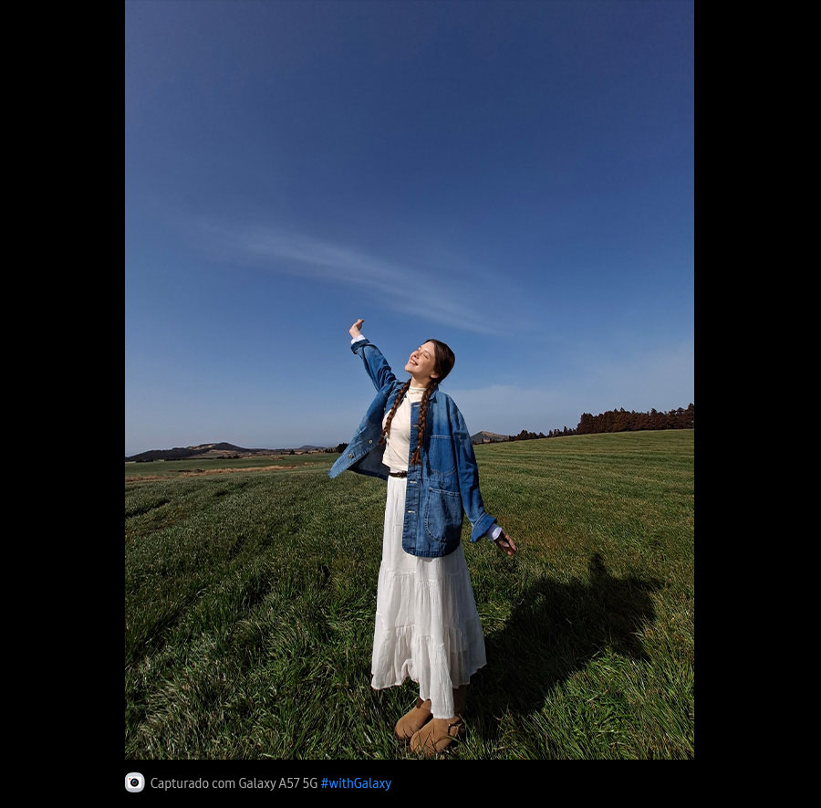 Foto de uma jovem em pé no meio de um campo gramado aberto sob um céu azul, capturada com a Câmera Ultra-Wide do Galaxy A57 5G. Os detalhes da imagem são claros e vívidos, com qualidade aprimorada. Na parte inferior, aparece um ícone de câmera com o texto 'Capturado com Galaxy A57 5G' e a hashtag #withGalaxy.