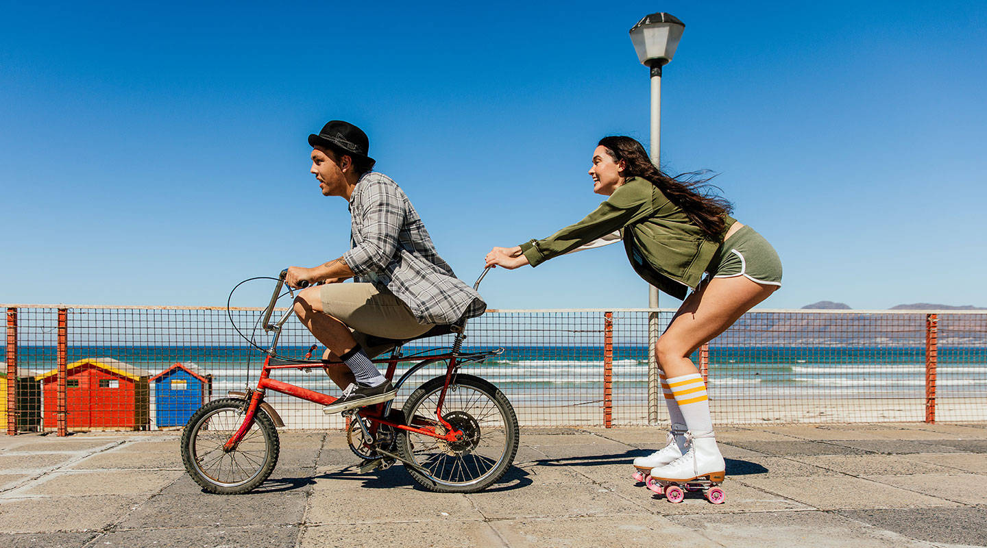  Uma imagem dividida ao meio mostra um homem e uma mulher próximos à praia. No lado esquerdo, a imagem parece levemente borrada, com o homem andando de bicicleta. No lado direito, a imagem está nítida e clara, mostrando a mulher segurando a bicicleta do homem enquanto usa patins, destacando o OIS (Estabilização Óptica de Imagem) do Galaxy A26 5G.
