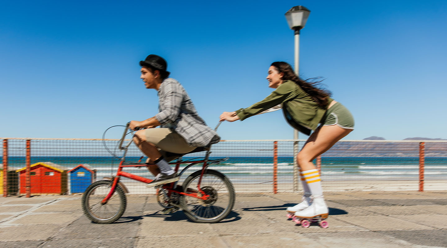  Uma imagem dividida ao meio mostra um homem e uma mulher próximos à praia. No lado esquerdo, a imagem parece levemente borrada, com o homem andando de bicicleta. No lado direito, a imagem está nítida e clara, mostrando a mulher segurando a bicicleta do homem enquanto usa patins, destacando o OIS (Estabilização Óptica de Imagem) do Galaxy A26 5G.