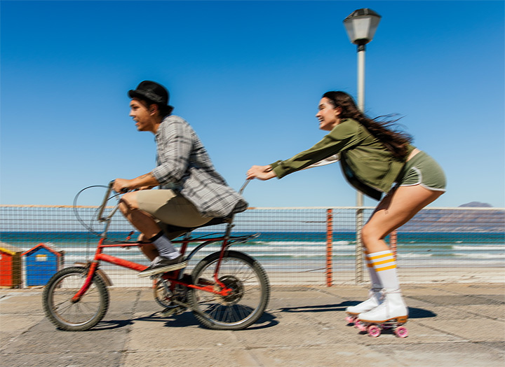  Uma imagem dividida ao meio mostra um homem e uma mulher próximos à praia. No lado esquerdo, a imagem parece levemente borrada, com o homem andando de bicicleta. No lado direito, a imagem está nítida e clara, mostrando a mulher segurando a bicicleta do homem enquanto usa patins, destacando o OIS (Estabilização Óptica de Imagem) do Galaxy A26 5G.