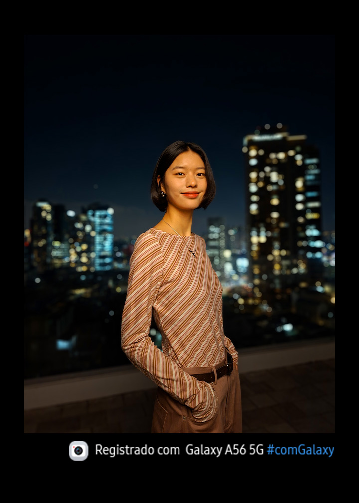 Vemos uma pessoa sorrindo para a c&acirc;mera e a vista noturna de uma cidade ao fundo. Na parte inferior, h&aacute; um &iacute;cone de c&acirc;mera com o texto "Capturado com Galaxy A56 5G #withGalaxy".