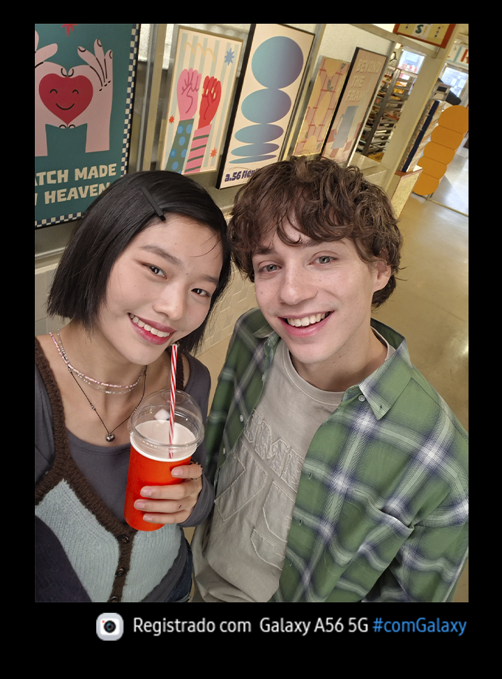 Vemos duas pessoas sorrindo em um cen&aacute;rio com fundo casual. A pessoa &agrave; esquerda est&aacute; segurando um copo de caf&eacute; descart&aacute;vel. O &acirc;ngulo indica que est&atilde;o tirando uma selfie. Na parte inferior, h&aacute; um &iacute;cone de c&acirc;mera com o texto "Capturado com Galaxy A56 5G #withGalaxy"