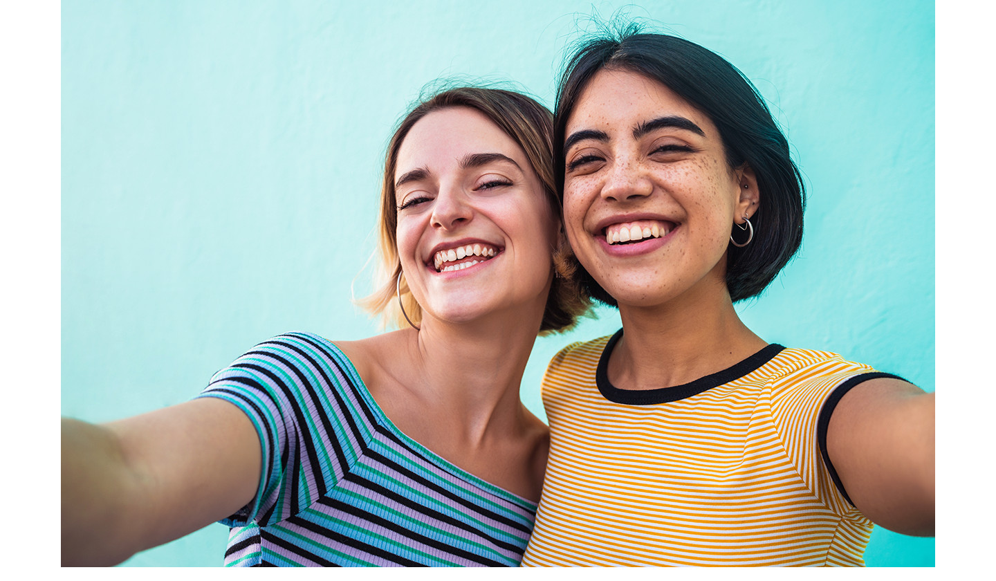 Duas mulheres sorriem enquanto tiram uma selfie juntas usando a Câmera Frontal de 12 MP do Galaxy A27 5G. A selfie tem cores vibrantes, detalhes nítidos e exposição equilibrada, com aparência natural.